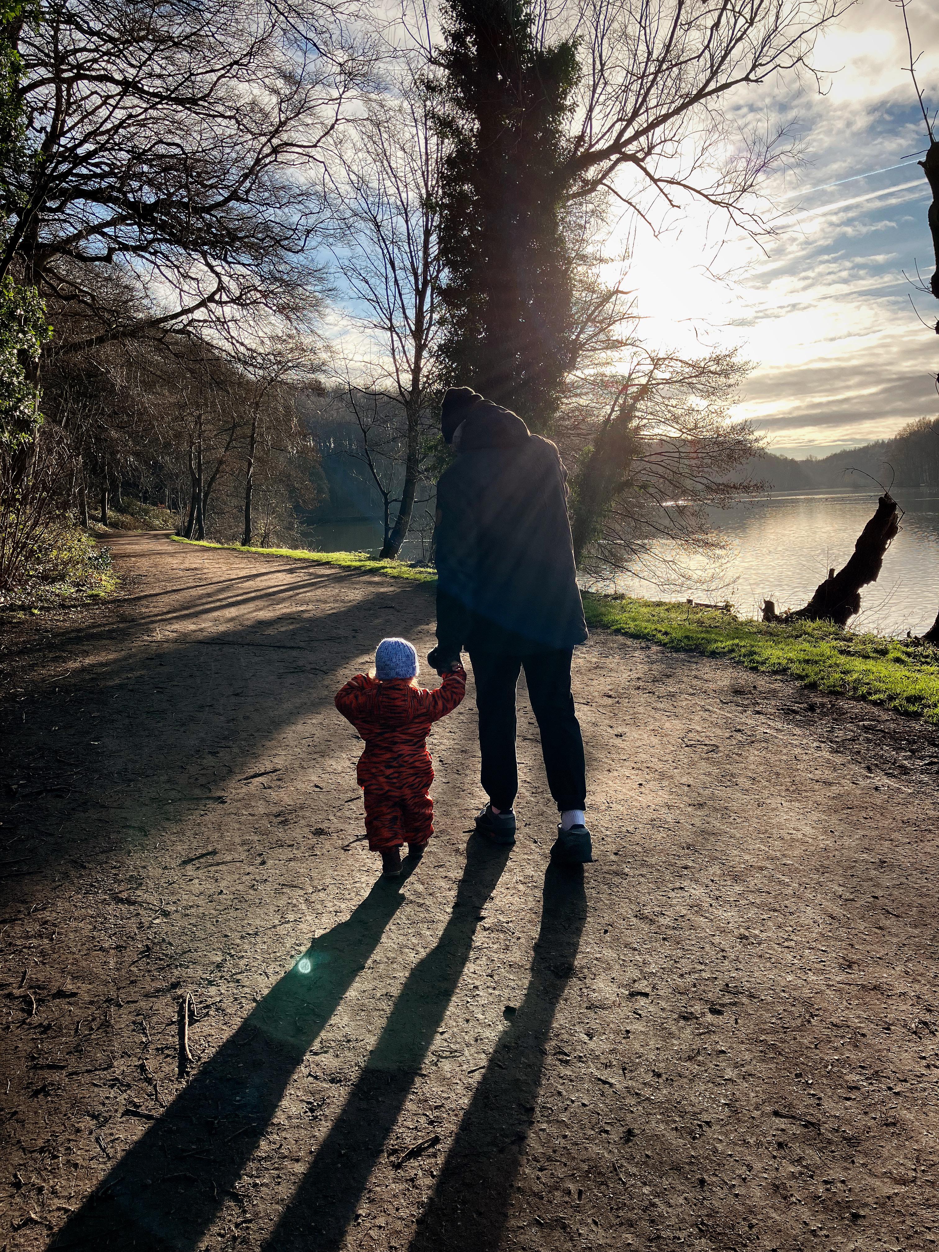 a toddler and his dad are walking away from the camera hand in hand. They are walking alongside a huge lake in the sunshine
